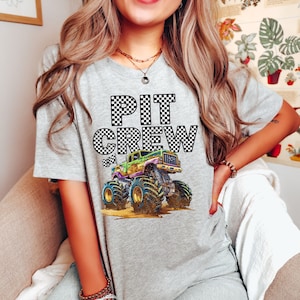 May include: A light gray t-shirt featuring the words "PIT CREW" in a checkered pattern. Below the text is a colorful monster truck illustration with large tires. The shirt is a casual style.