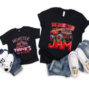 Two black T-shirts one child sized says "MONSTER TRUCKS ARE MY JAM" in pink and strawberry letters with a red monster truck. The adult sized one says "MOMSTER JAM" in strawberry letters with a red monster rtuck
