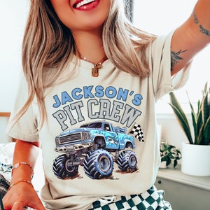 Pictured is a woman wearing a Cream-colored t-shirt featuring a blue shark themed monster truck graphic with the text "JACKSON'S PIT CREW". The truck has large tires and a checkered flag. The shirt is a crew neck and is perfect as a complimentary tee for boys birthday party
