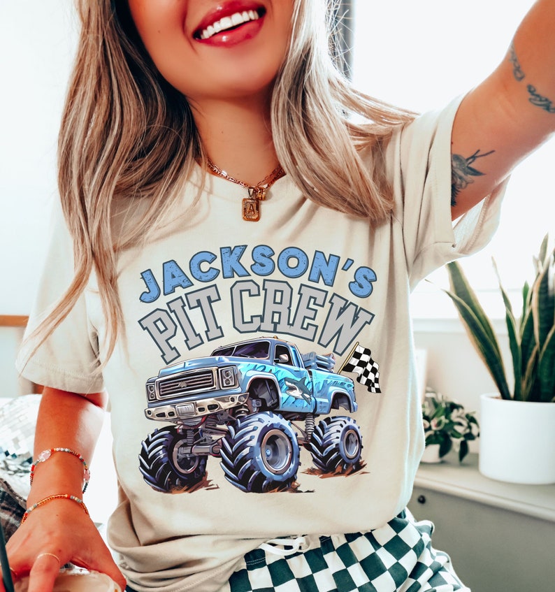 Pictured is a woman wearing a Cream-colored t-shirt featuring a blue shark themed monster truck graphic with the text "JACKSON'S PIT CREW". The truck has large tires and a checkered flag. The shirt is a crew neck and is perfect as a complimentary tee for boys birthday party