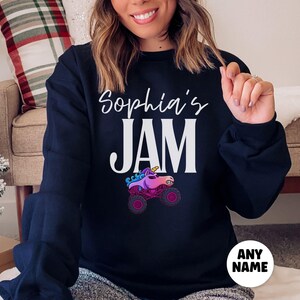 May include: A navy blue sweatshirt with the text "Sophia's JAM" in white script and block letters. Below the text is a graphic of a pink and purple unicorn monster truck. A white circle with the text "ANY NAME" is at the bottom.