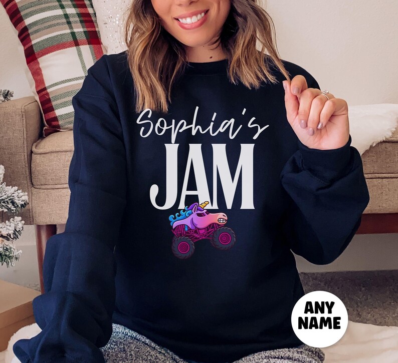 May include: A navy blue sweatshirt with the text "Sophia's JAM" in white script and block letters. Below the text is a graphic of a pink and purple unicorn monster truck. A white circle with the text "ANY NAME" is at the bottom.
