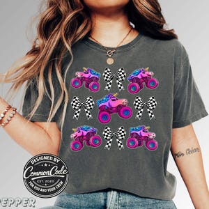 May include: Dark grey t-shirt with a vibrant graphic of monster trucks with unicorn heads and checkered bows. The design incorporates pink, purple, blue, and black hues. The shirt has a crew neck and short sleeves. The text "DESIGNED BY COMMON CODE EST. 2020 FOR YOU AND YOUR CREW" is printed on the shirt.