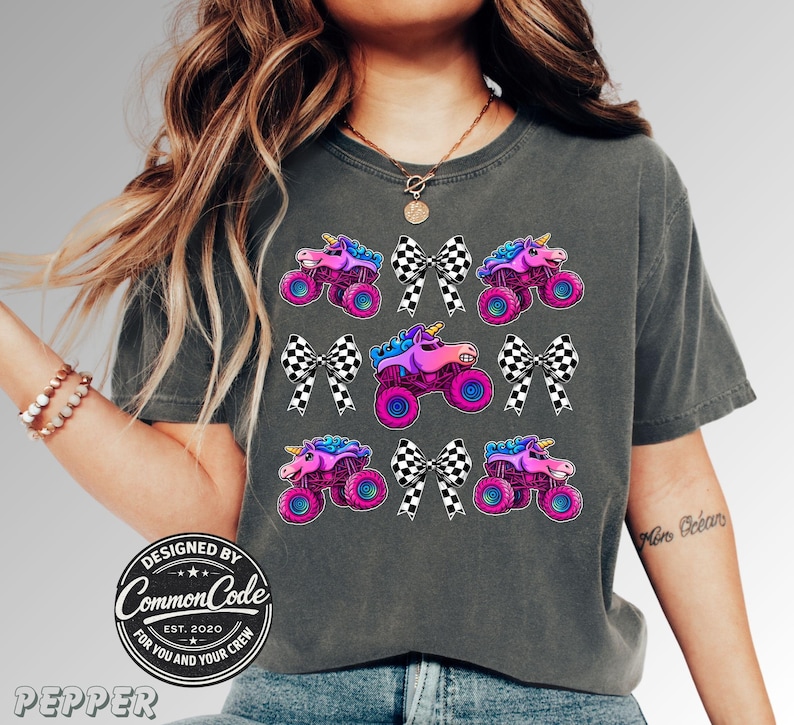 May include: Dark grey t-shirt with a vibrant graphic of monster trucks with unicorn heads and checkered bows. The design incorporates pink, purple, blue, and black hues. The shirt has a crew neck and short sleeves. The text "DESIGNED BY COMMON CODE EST. 2020 FOR YOU AND YOUR CREW" is printed on the shirt.