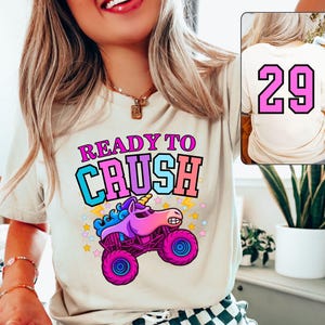 Pode incluir: Uma camiseta creme com o texto "READY TO CRUSH" em rosa, azul e laranja. Um desenho animado de um caminh&atilde;o monstro unic&oacute;rnio est&aacute; abaixo do texto. A parte de tr&aacute;s da camiseta tem o n&uacute;mero "29" em rosa.