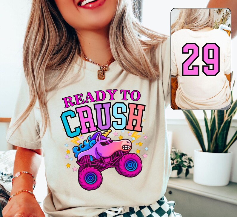 Pode incluir: Uma camiseta creme com o texto "READY TO CRUSH" em rosa, azul e laranja. Um desenho animado de um caminh&atilde;o monstro unic&oacute;rnio est&aacute; abaixo do texto. A parte de tr&aacute;s da camiseta tem o n&uacute;mero "29" em rosa.