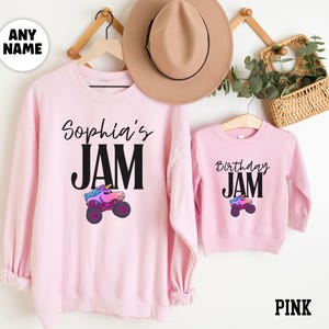 May include: Pink sweatshirts with the text "Sophia's JAM" and "Birthday JAM". Each sweatshirt features a colorful monster truck graphic. A tan hat and a small basket with greenery are also visible.