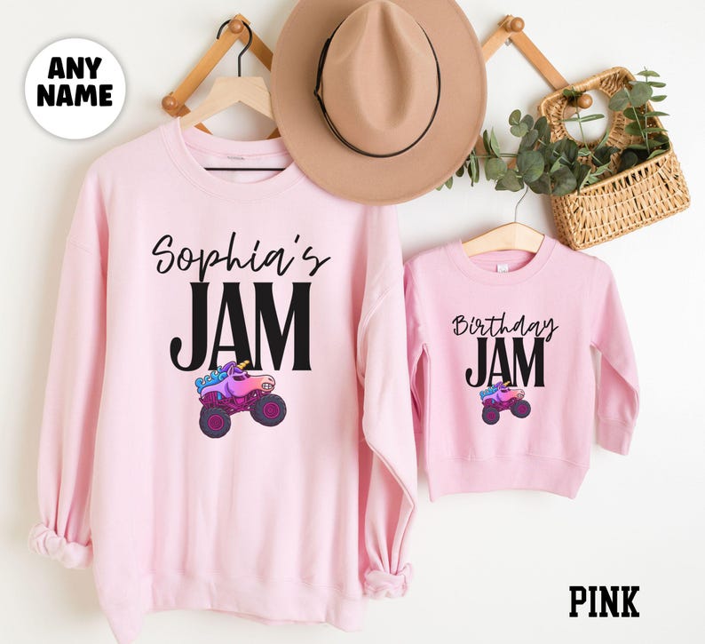 May include: Pink sweatshirts with the text "Sophia's JAM" and "Birthday JAM". Each sweatshirt features a colorful monster truck graphic. A tan hat and a small basket with greenery are also visible.