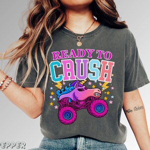 May include: Dark gray t-shirt with a graphic of a monster truck unicorn and the text "READY TO CRUSH". The design features a pink, purple, and blue unicorn on a monster truck with stars and lightning bolts.