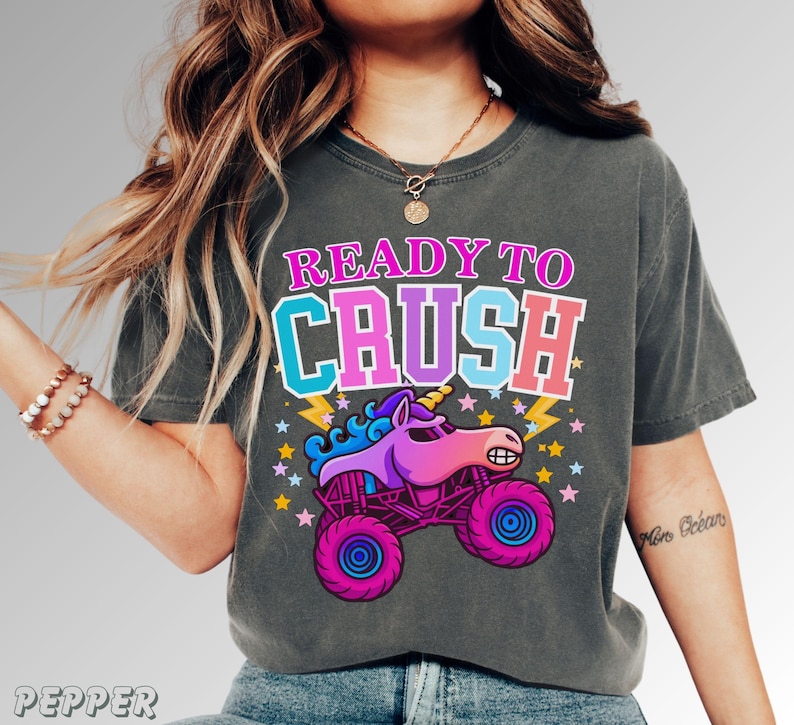 May include: Dark gray t-shirt with a graphic of a monster truck unicorn and the text "READY TO CRUSH". The design features a pink, purple, and blue unicorn on a monster truck with stars and lightning bolts.