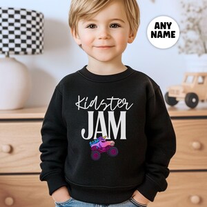 May include: Black crewneck sweatshirt with the text "Kidster JAM" in white script. A colorful graphic of a monster truck with a unicorn horn is below the text. A white circle with the text "ANY NAME" is in the upper right corner.