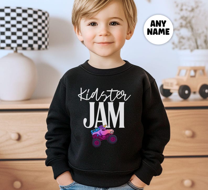 May include: Black crewneck sweatshirt with the text "Kidster JAM" in white script. A colorful graphic of a monster truck with a unicorn horn is below the text. A white circle with the text "ANY NAME" is in the upper right corner.