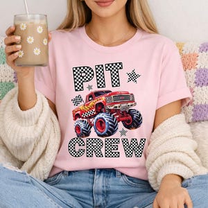 Op de afbeelding: Roze T-shirt met de tekst "PIT CREW" in een dambordpatroon. Een rode monstertruck-afbeelding staat in het midden. Het shirt heeft korte mouwen en een ronde hals.