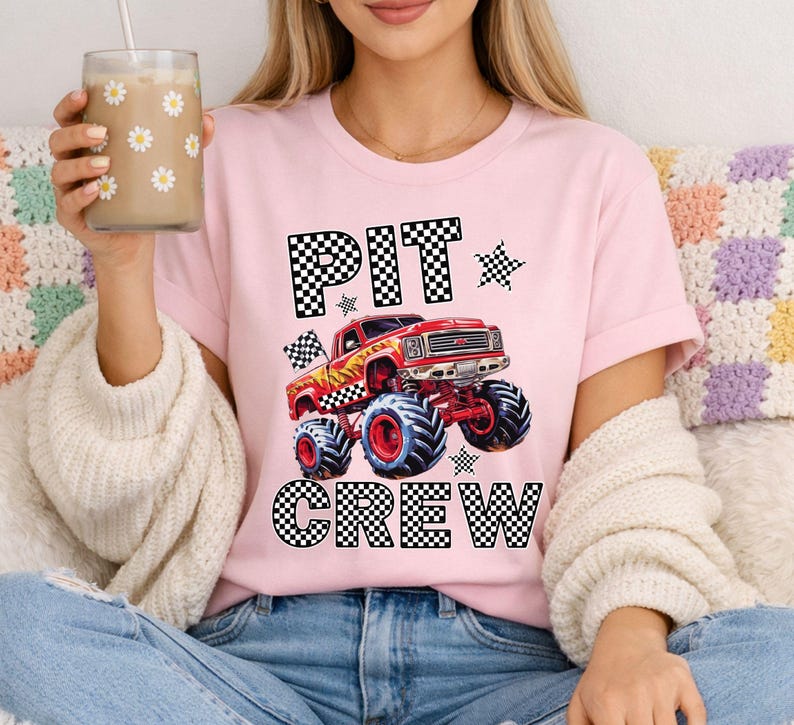 Op de afbeelding: Roze T-shirt met de tekst "PIT CREW" in een dambordpatroon. Een rode monstertruck-afbeelding staat in het midden. Het shirt heeft korte mouwen en een ronde hals.