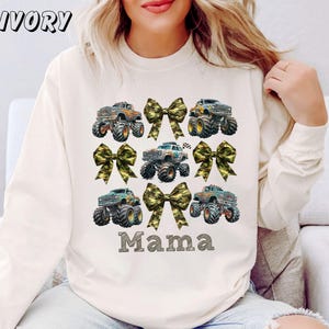 May include: Ivory-colored sweatshirt featuring a design of monster trucks and camouflage bows. The word "Mama" is printed in a block letter font below the trucks. The word "IVORY" is printed in the upper left corner.