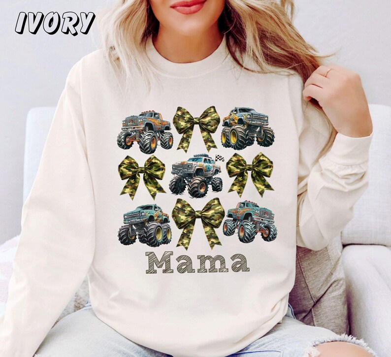 May include: Ivory-colored sweatshirt featuring a design of monster trucks and camouflage bows. The word "Mama" is printed in a block letter font below the trucks. The word "IVORY" is printed in the upper left corner.