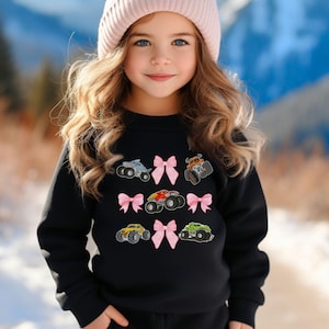 May include: Black sweatshirt featuring embroidered monster trucks in various colors, including gray, orange, red, yellow, and green. Pink bows are interspersed between the trucks. The sweatshirt is a children's apparel item.