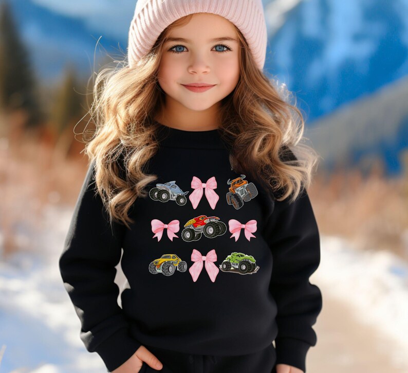 May include: Black sweatshirt featuring embroidered monster trucks in various colors, including gray, orange, red, yellow, and green. Pink bows are interspersed between the trucks. The sweatshirt is a children's apparel item.