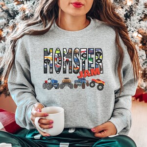 May include: Gray sweatshirt with the text "MOMSTER JAM" in colorful letters. The letters are made up of images of monster trucks.