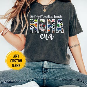 Puede incluir: Una camiseta gris oscuro con un gr&aacute;fico de la palabra "MAMA" formada por camiones monstruo. El texto "in my monster truck era" est&aacute; por encima del gr&aacute;fico. La camiseta la lleva puesta una persona con un tatuaje en el brazo.