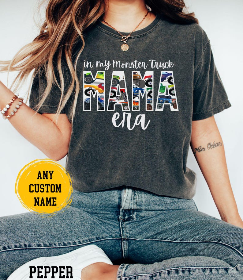 Puede incluir: Una camiseta gris oscuro con un gr&aacute;fico de la palabra "MAMA" formada por camiones monstruo. El texto "in my monster truck era" est&aacute; por encima del gr&aacute;fico. La camiseta la lleva puesta una persona con un tatuaje en el brazo.