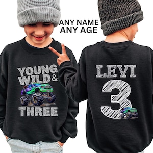 Op de afbeelding: Zwart sweatshirt met een monstertruck-afbeelding en de tekst "Young Wild & Three". De achterkant van het sweatshirt heeft een naam en nummer in een wit, gekrabbeld lettertype.