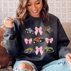 May include: Dark gray sweatshirt featuring colorful monster truck graphics and pink bows. The design includes a blue, orange, red, yellow, and green monster truck. The sweatshirt is a comfortable, casual style.