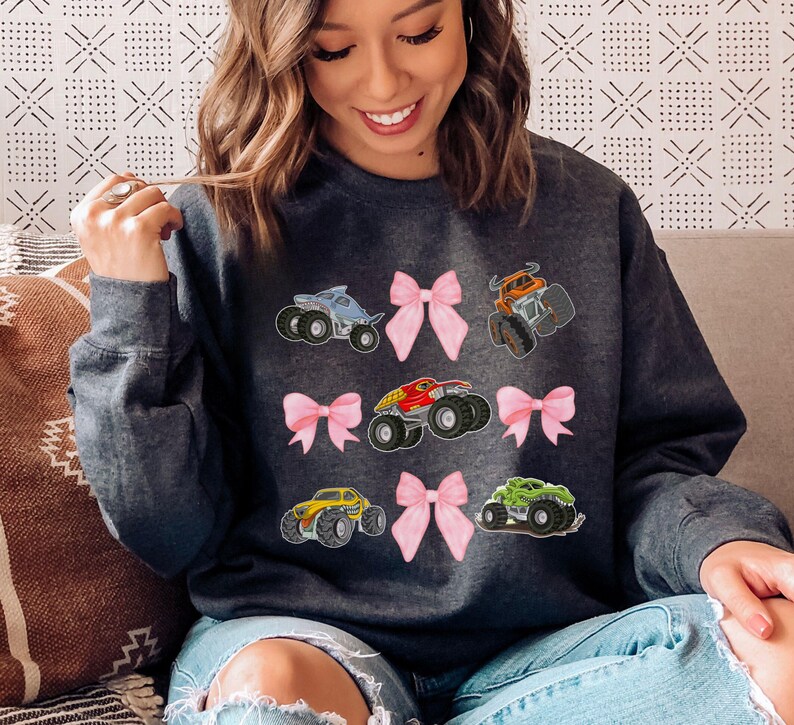 May include: Dark gray sweatshirt featuring colorful monster truck graphics and pink bows. The design includes a blue, orange, red, yellow, and green monster truck. The sweatshirt is a comfortable, casual style.