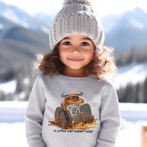 Op de afbeelding: Lichtgrijs sweatshirt met een oranje monstertruck ontwerp, grote banden en modderspatten. De tekst "A LITTLE DIRT DOESN'T HURT" staat eronder. Het sweatshirt wordt gedragen door een kind met een grijze gebreide muts.