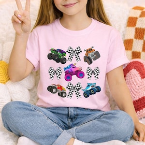 May include: A light pink t-shirt featuring colorful monster truck graphics with checkered bows. The monster trucks have various designs, including a shark, a unicorn, and a truck with flames. The shirt is paired with blue jeans.