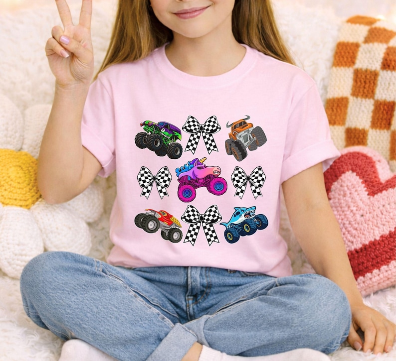 May include: A light pink t-shirt featuring colorful monster truck graphics with checkered bows. The monster trucks have various designs, including a shark, a unicorn, and a truck with flames. The shirt is paired with blue jeans.