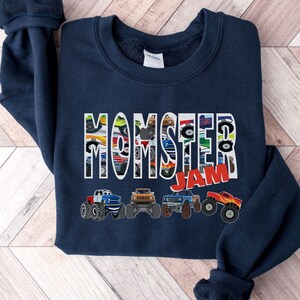 May include: Navy blue sweatshirt with the words "Monster Jam" printed in white. The letters are made up of images of monster trucks. There are five monster trucks printed below the text.