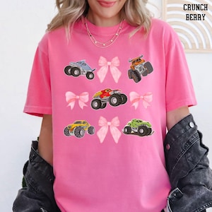 Pictured is a woman wearing a crunch berry pink t-shirt which has 4 pink coquette bows and 5 monster trucks in a grid tic tac toe pattern