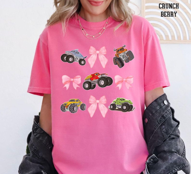 Pictured is a woman wearing a crunch berry pink t-shirt which has 4 pink coquette bows and 5 monster trucks in a grid tic tac toe pattern