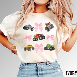 K&ouml;nnte beinhalten: Elfenbeinfarbenes T-Shirt mit farbenfrohen Monstertruck-Grafiken und rosa Schleifen. Die Trucks sind in verschiedenen Farben gehalten, darunter Blau, Orange, Rot, Gelb und Gr&uuml;n. Das Wort "IVORY" ist unten in Schwarz gedruckt.