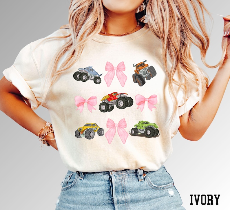K&ouml;nnte beinhalten: Elfenbeinfarbenes T-Shirt mit farbenfrohen Monstertruck-Grafiken und rosa Schleifen. Die Trucks sind in verschiedenen Farben gehalten, darunter Blau, Orange, Rot, Gelb und Gr&uuml;n. Das Wort "IVORY" ist unten in Schwarz gedruckt.