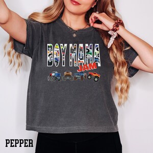 K&ouml;nnte beinhalten: Ein dunkelgraues T-Shirt mit dem Aufdruck "Boy Mama Jam" in bunten Buchstaben. Die Buchstaben bestehen aus Bildern von Monstertrucks. Unter dem Text befinden sich f&uuml;nf Monstertrucks in einer Reihe.