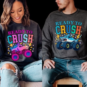 Pu&ograve; includere: Due felpe grigio scuro con la scritta "READY TO CRUSH". Una felpa presenta un monster truck unicorno, l'altra un monster truck squalo. Entrambi i design hanno accenti colorati e stelle.