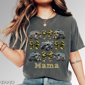 May include: Dark gray t-shirt featuring a design of monster trucks and camouflage bows. The word "Mama" is printed in gold block letters. The shirt has a crew neck and short sleeves.
