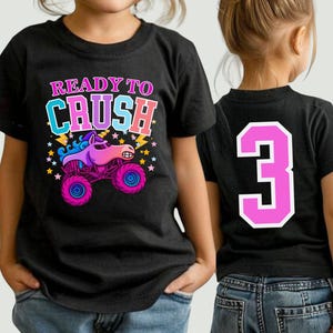 Pode incluir: T-shirt preto com a frase "READY TO CRUSH" e um desenho animado de um monster truck com cabe&ccedil;a de unic&oacute;rnio. O caminh&atilde;o &eacute; rosa e roxo com rodas azuis. O n&uacute;mero "3" est&aacute; nas costas da camisa.