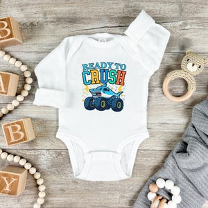 Pu&ograve; includere: Body da bambino bianco a maniche lunghe. Presenta una grafica di uno squalo su ruote di monster truck con la scritta "READY TO CRUSH" in blu, arancione e giallo. Blocchi di legno e un orsacchiotto sullo sfondo.