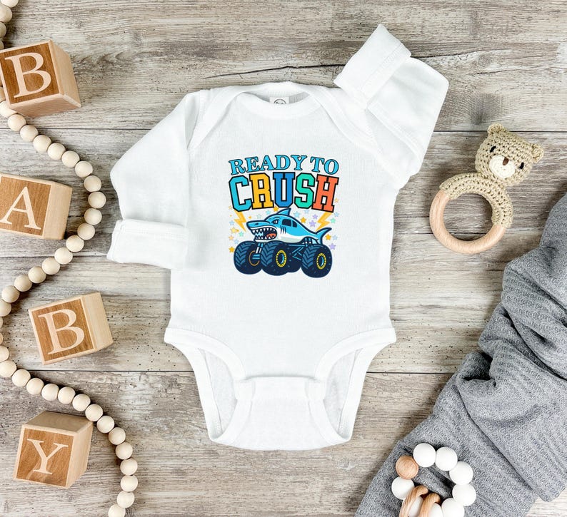 Pu&ograve; includere: Body da bambino bianco a maniche lunghe. Presenta una grafica di uno squalo su ruote di monster truck con la scritta "READY TO CRUSH" in blu, arancione e giallo. Blocchi di legno e un orsacchiotto sullo sfondo.