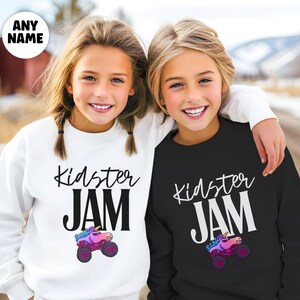 May include: Two children wearing matching sweatshirts. One sweatshirt is white, the other is black. Both sweatshirts have the words "Kidster JAM" in black and white, with a cartoon unicorn monster truck graphic.