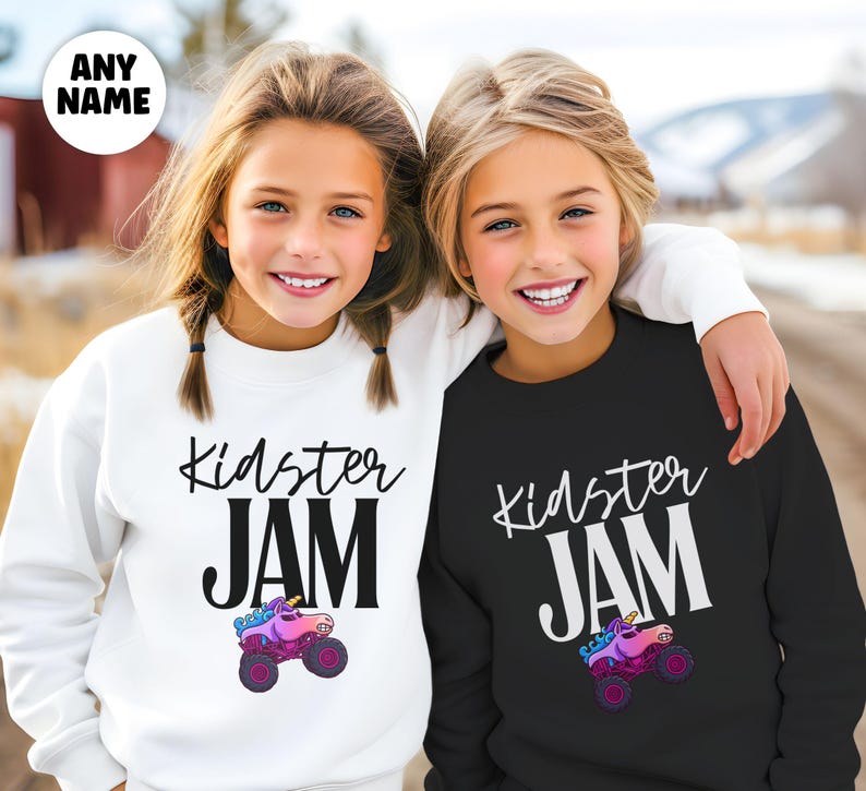 May include: Two children wearing matching sweatshirts. One sweatshirt is white, the other is black. Both sweatshirts have the words "Kidster JAM" in black and white, with a cartoon unicorn monster truck graphic.