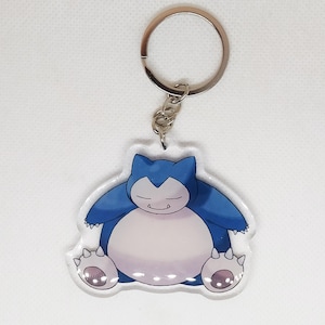 Può includere: Un portachiavi Snorlax blu e bianco. Il portachiavi presenta un'immagine a cartoni animati di Snorlax, un grande e sonnolento Pokémon, con un corpo azzurro, dettagli blu scuro e un portachiavi argentato.