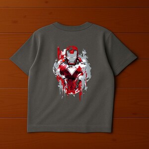 Puede incluir: Camiseta gris con un gr&aacute;fico de Iron Man en rojo, plateado y blanco. El dise&ntilde;o tiene un efecto de salpicaduras de pintura desgastadas. El casco y la armadura del superh&eacute;roe se muestran prominentemente en la parte delantera de la camiseta.