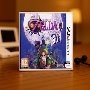 Puede incluir: Caja del juego The Legend of Zelda: Majora's Mask 3D para Nintendo 3DS. La portada presenta a Link, una gran luna y varios personajes. La caja tiene un borde blanco y el logotipo de Nintendo 3DS.
