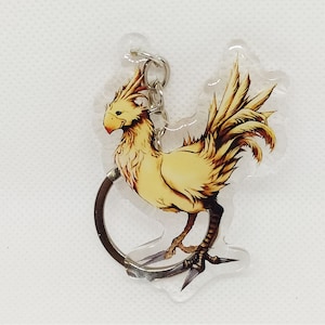 May include: A clear acrylic keychain featuring a yellow Chocobo bird character with a red beak and detailed feathering. The keychain has a silver metal ring for attaching to keys or bags. The Chocobo is a popular character from the Final Fantasy video game series.