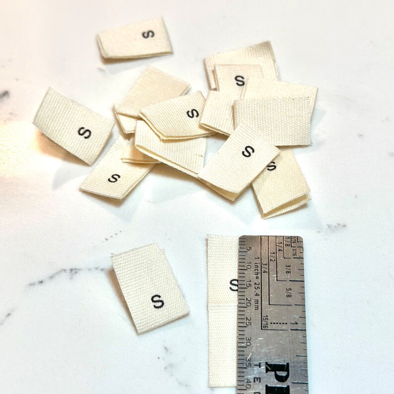 May include: A pile of off-white fabric labels with the letter "S" printed in black. A ruler is in the bottom right corner of the image.