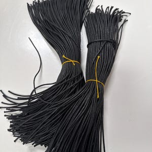 May include: Two bundles of thin, black, round cord, each secured in the center with a bright yellow tie. The cords are arranged in parallel, straight lines, creating a dense, uniform texture of black strands against a plain white background.